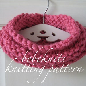 May include: A pink knitted cowl neck scarf hanging on a white wooden hanger. The scarf is made with a chunky yarn and has a simple, textured stitch pattern. The text "bebeknits knitting pattern" is written below the scarf.