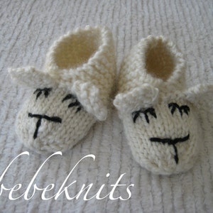 May include: A pair of white knitted baby booties with a sheep face design. The booties have black embroidered eyes, nose, and mouth.