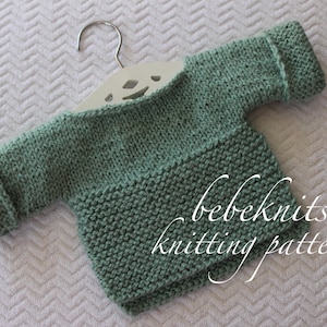Puede incluir: Un jersey de punto verde para bebé con textura de canalé, colgado de una percha blanca. El jersey tiene un bolsillo en la parte delantera y puños enrollados. El texto "bebeknits knitting pattern" es visible en el jersey.