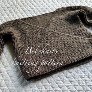 May include: A brown knitted baby cardigan with a wrap-style front and two brown buttons. The cardigan is laid flat on a white quilted surface. The text "Bebeknits knitting pattern" is visible on the cardigan.