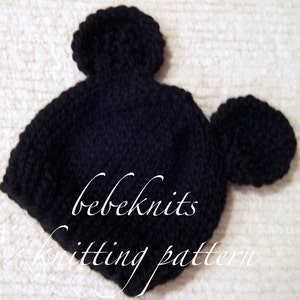 May include: Black knitted baby hat with two large pom-poms on top, resembling Mickey Mouse ears. The hat has the text "bebeknits knitting pattern" on the front.