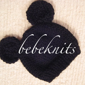 May include: Black knitted baby hat with two large pom-poms on top. The hat has the text "bebeknits" written on it.