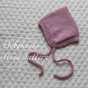May include: A knitted bonnet in a soft mauve color, featuring a square crown and ties. The bonnet is displayed on a white, quilted surface. The text "Bebeknits knitting pattern" is visible.