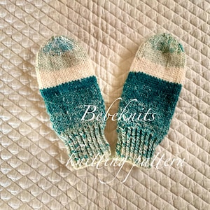 May include: A pair of knitted baby mittens in a green, white, and cream color gradient. The mittens are made with a soft, textured yarn and have a simple, classic design.