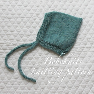 以下が含まれることがあります： 紐付きのターコイズブルーのニットのベビーボンネット。ボンネットは白いキルティングの表面の上に置かれています。ボンネットの下に「Bebeknits knitting pattern」というテキストが表示されています。
