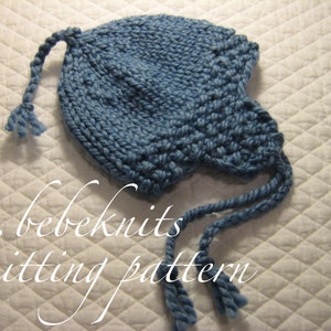 May include: A blue knitted baby hat with two long ties. The hat has a pointed top and a brim that covers the ears. The ties are tied together at the back of the hat. The hat is on a white quilted surface. bebeknits knitting pattern.