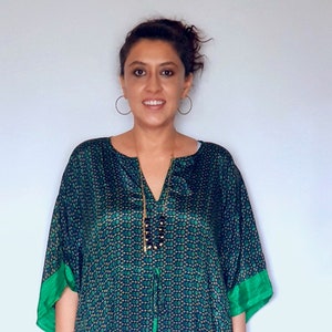 May include: A woman wearing a long, green and blue patterned kaftan with a green trim. The kaftan has a V-neck and a tie at the neckline. She is wearing a silver necklace and silver earrings. She is also wearing a silver bracelet on her right wrist.