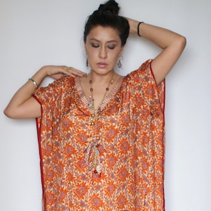 Op de afbeelding: Een lange, oranje en witte gepatroneerde kaftan met een rode zoom. De kaftan heeft een V-hals en is gemaakt van een zijdeachtige stof. Het model draagt de kaftan met een zilveren armband en een lange kralenketting.
