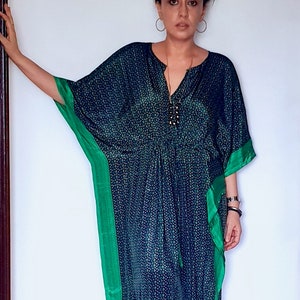 May include: A woman wearing a blue and green patterned kaftan with a green trim. She is wearing a gold necklace with a black pendant.