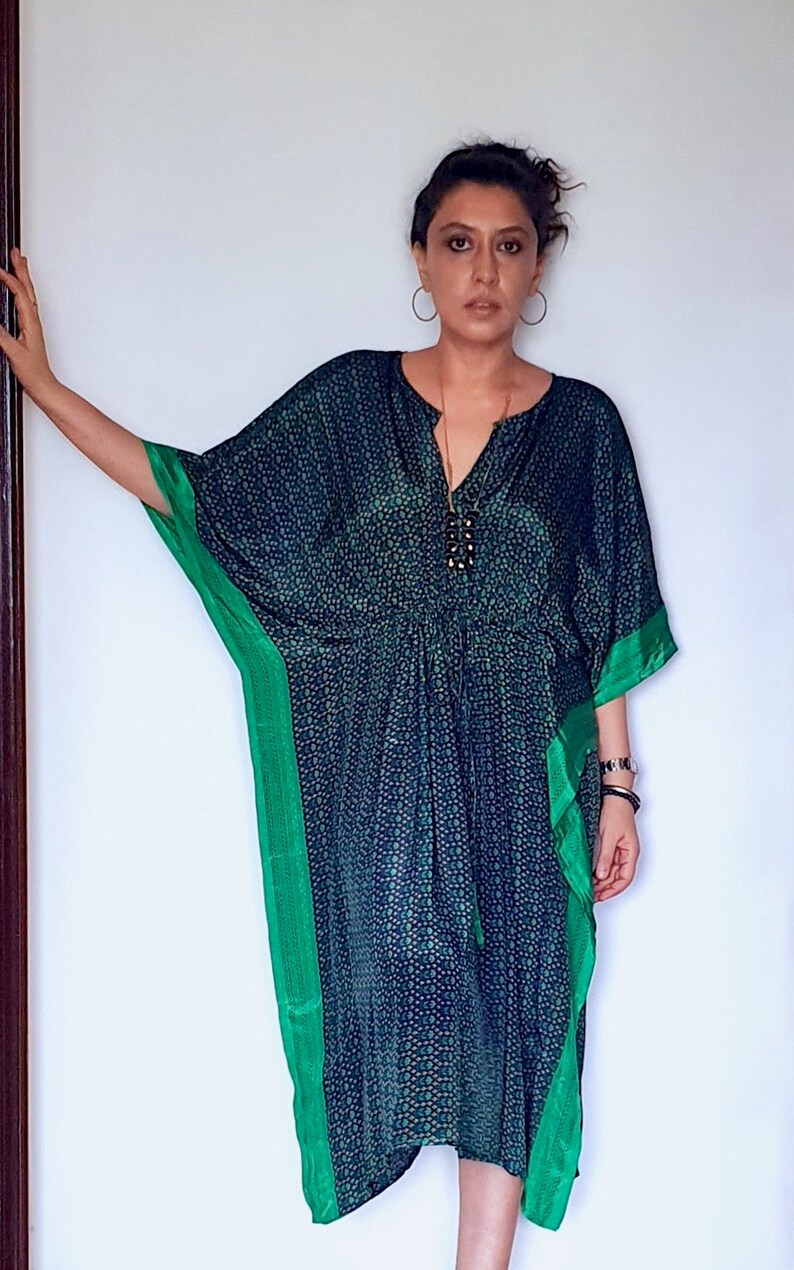 May include: A woman wearing a blue and green patterned kaftan with a green trim. She is wearing a gold necklace with a black pendant.