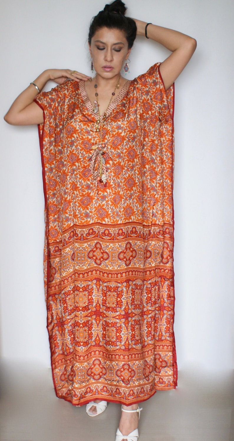 Op de afbeelding: Een lange, oranje en witte gepatroneerde kaftan met een rode zoom. De kaftan heeft een V-hals en is gemaakt van een zijdeachtige stof. Het model draagt de kaftan met een zilveren armband en een lange kralenketting.