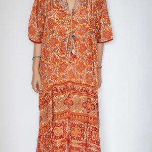 Op de afbeelding: Een lange oranje en witte kaftan met een bloemenprint, een V-hals en 3/4 mouwen. De kaftan heeft een strik in de taille en een kwastdetail.