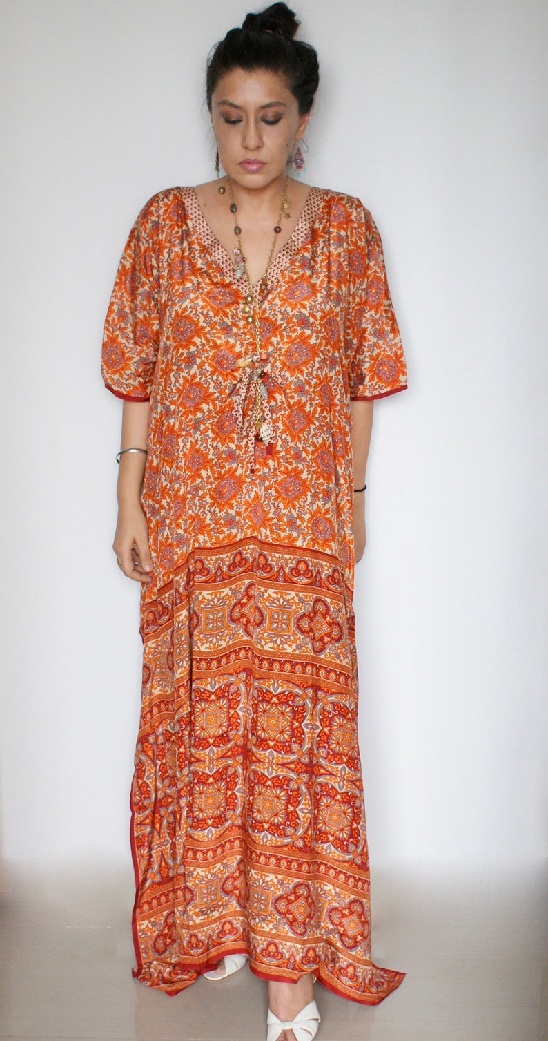 Op de afbeelding: Een lange oranje en witte kaftan met een bloemenprint, een V-hals en 3/4 mouwen. De kaftan heeft een strik in de taille en een kwastdetail.