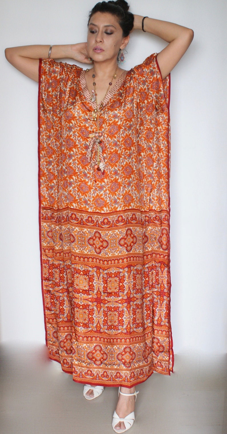 Op de afbeelding: Een lange, vloeiende oranje en rode gepatroneerde kaftan met een V-hals. De kaftan is gemaakt van een zachte, zijdeachtige stof en heeft een ontspannen pasvorm. Het model draagt de kaftan met een zilveren armband en een lange kralenketting.
