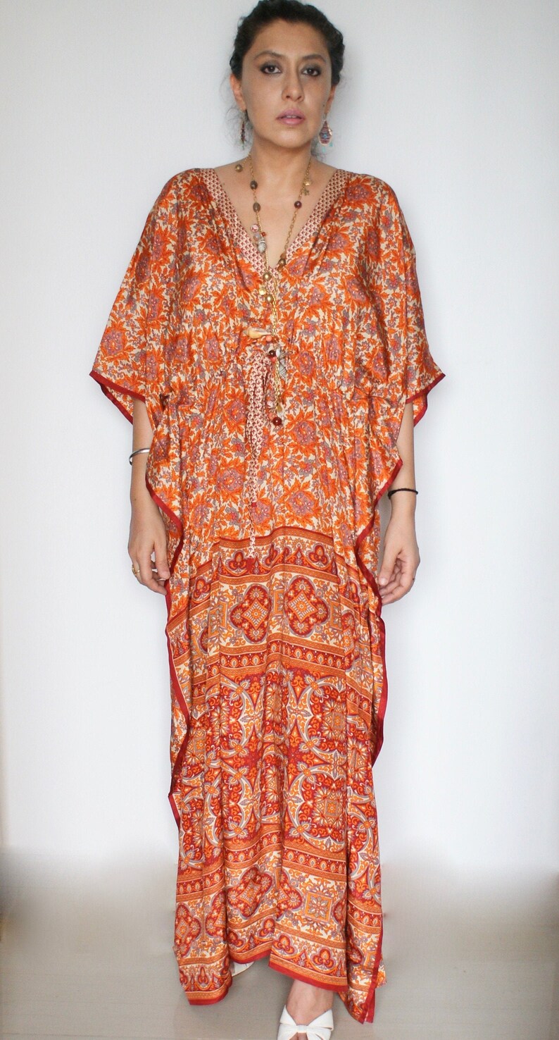 Op de afbeelding: Een lange, vloeiende oranje en rode gepatroneerde kaftan met een V-hals en een strik in de taille. De kaftan is gemaakt van een zachte, zijdeachtige stof en heeft een ontspannen pasvorm.