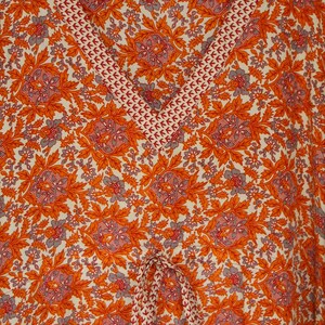 Op de afbeelding: Een witte, oranje en grijze stof met een bloemenprint en een rode en witte bies. De stof heeft een trekkoord.
