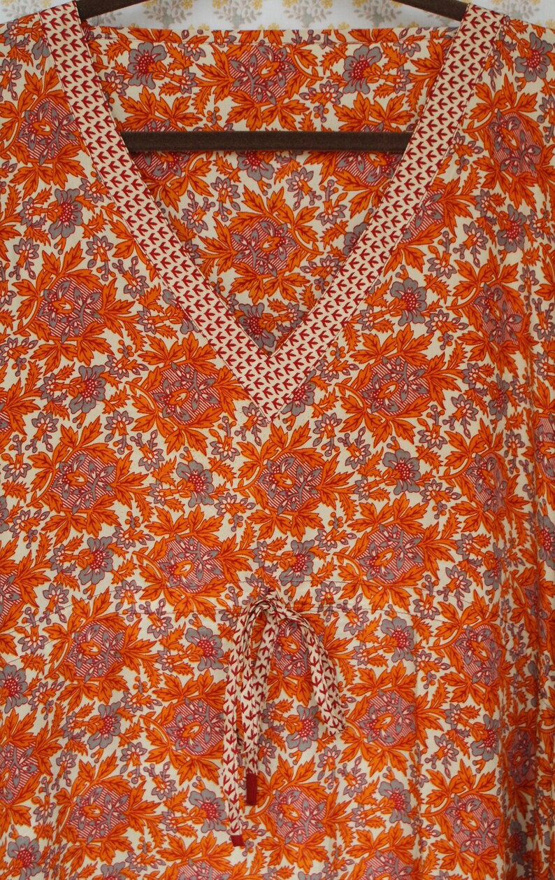 Op de afbeelding: Een witte, oranje en grijze stof met een bloemenprint en een rode en witte bies. De stof heeft een trekkoord.