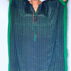 May include: A woman wearing a blue and green patterned kaftan with a green trim. The kaftan has a V-neck and a long, flowing silhouette.