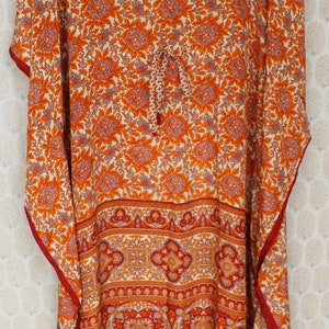 Op de afbeelding: Een lange, vloeiende oranje en witte geprinte kaftan met een V-hals en een strik in de taille. De kaftan heeft een rode bies en een geometrisch patroon aan de onderkant.