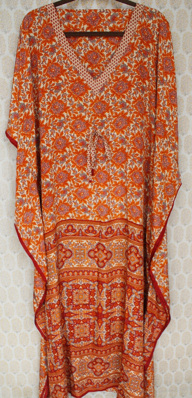 Op de afbeelding: Een lange, vloeiende oranje en witte geprinte kaftan met een V-hals en een strik in de taille. De kaftan heeft een rode bies en een geometrisch patroon aan de onderkant.