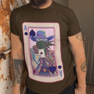 以下が含まれることがあります： ダークブラウンの半袖Tシャツで、スペードのエースのトランプカードの大きなグラフィックプリントが施されています。帽子とバンダナを身に着け、トランプカードを1枚持ったカウボーイの姿が描かれており、ピンクの枠で囲まれています。