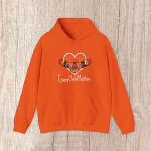 May include: Bright orange hoodie with a front pocket and a drawstring hood. The design features a white heart with three figures and the text "Every Child Matters" in white.