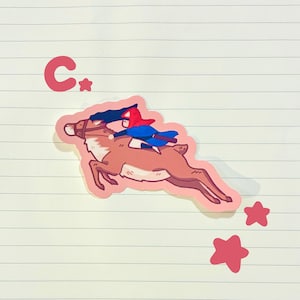 May include: A sticker featuring a cartoon reindeer with a rider in a red hood and blue cape. The reindeer is brown with white accents, and the rider is holding a black object. Pink stars and the letter "C" with a star are also included.