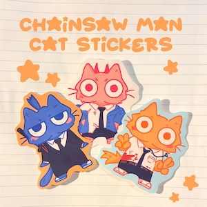 May include: Three die-cut stickers featuring cartoon cats dressed in suits, with the text "CHAINSAW MAN CAT STICKERS" in orange. One cat is blue, one is pink, and one is orange, all with large eyes and stylized outfits on lined paper.
