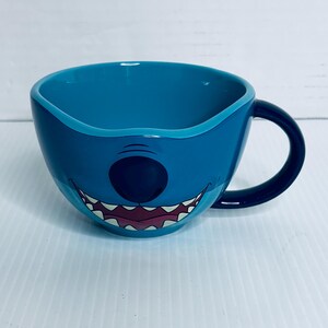 K&ouml;nnte beinhalten: Eine breite, runde Keramiktasse in leuchtendem Blau mit einem grafischen Aufdruck eines Charaktergesichts, das eine gro&szlig;e schwarze Nase und ein breites, zahniges Grinsen zeigt. Die Tasse hat eine glatte, gl&auml;nzende Oberfl&auml;che und einen geschwungenen Henkel.
