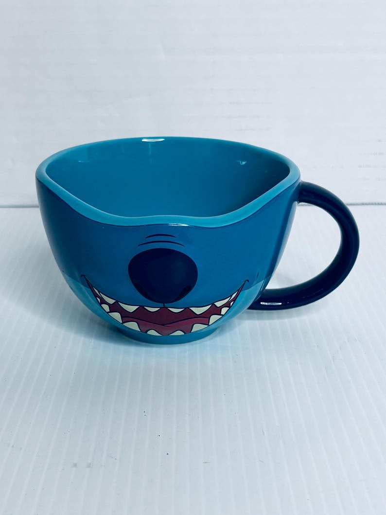 K&ouml;nnte beinhalten: Eine breite, runde Keramiktasse in leuchtendem Blau mit einem grafischen Aufdruck eines Charaktergesichts, das eine gro&szlig;e schwarze Nase und ein breites, zahniges Grinsen zeigt. Die Tasse hat eine glatte, gl&auml;nzende Oberfl&auml;che und einen geschwungenen Henkel.