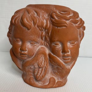 May include: A terracotta-colored ceramic planter featuring the sculpted faces of two cherubs side by side. The design includes detailed hair, facial features, and small wings positioned below each face, with a matte finish and white highlights.