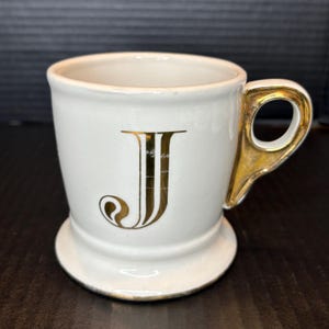 May include: White ceramic mug with a gold handle and a gold letter "J" on the front. The mug has a pedestal base and a gold rim. The mug is likely for beverages.