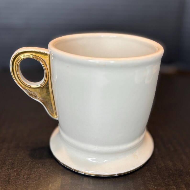 May include: A white ceramic mug with a gold-colored handle. The mug has a wide base and a slightly flared rim. The handle is a unique shape, adding a touch of elegance to the design. The mug is clean and simple.