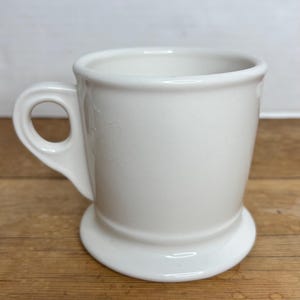 Pu&ograve; includere: Una tazza in ceramica bianca con base arrotondata e manico. La tazza ha un design semplice e classico ed &egrave; adatta per bevande calde. La tazza &egrave; su una superficie di legno.