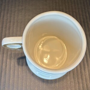 May include: An overhead view of a white ceramic mug with a handle. The interior of the mug is a light cream color. The mug sits on a small, circular base. The handle is a curved shape.