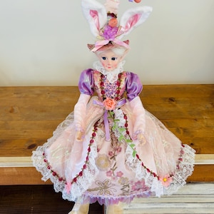 May include: A decorative Easter doll with a whimsical design. The doll features a pink and purple dress with lace trim, a matching hat with bunny ears, and a carrot accent. The doll has a painted face and is designed for Easter decor.