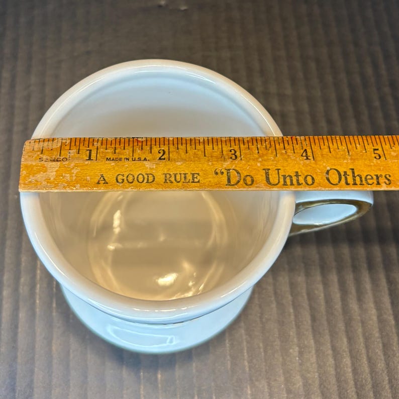 May include: A white ceramic mug with a gold handle and trim. A wooden ruler is placed across the top of the mug, measuring approximately 4 inches across. The ruler has the text "A GOOD RULE Do Unto Others".