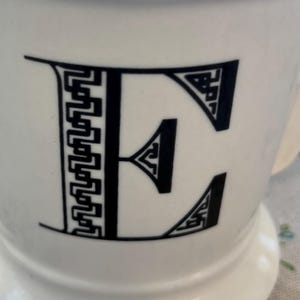 Puede incluir: Taza de cer&aacute;mica blanca con una letra "E" negra en una fuente decorativa y geom&eacute;trica. La taza tiene una base ancha y un asa curvada. El dise&ntilde;o es simple pero elegante, lo que la convierte en una adici&oacute;n vers&aacute;til a cualquier cocina.