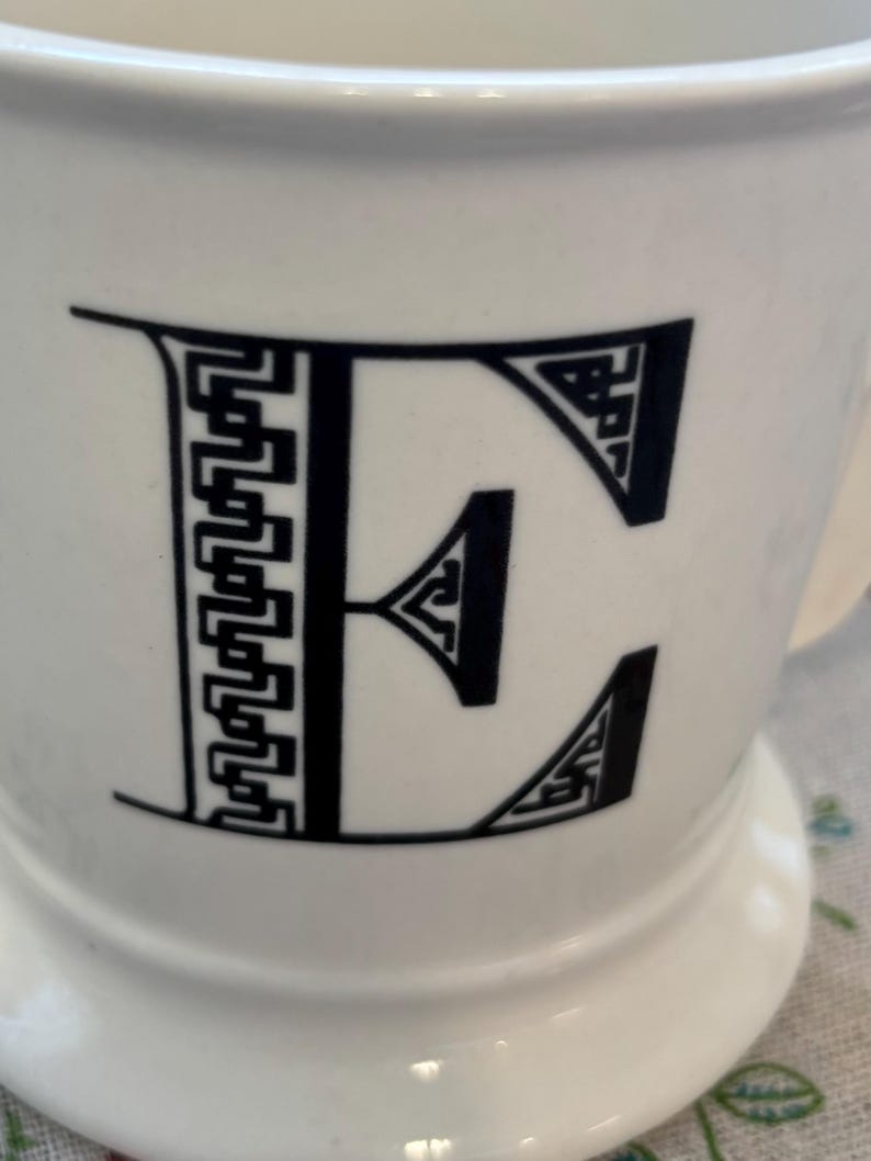 Puede incluir: Taza de cer&aacute;mica blanca con una letra "E" negra en una fuente decorativa y geom&eacute;trica. La taza tiene una base ancha y un asa curvada. El dise&ntilde;o es simple pero elegante, lo que la convierte en una adici&oacute;n vers&aacute;til a cualquier cocina.