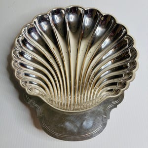 May include: A silver-colored, shell-shaped dish with a scalloped edge. The dish has a detailed, ribbed design radiating from the center. The base of the dish has a decorative, rectangular shape.