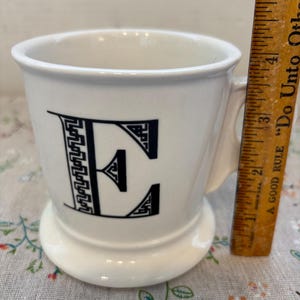 Puede incluir: Taza de cer&aacute;mica blanca con un dise&ntilde;o de la letra "E" en negro. La taza tiene un asa y una base ancha. Una regla de madera est&aacute; al lado de la taza, midiendo aproximadamente 10 cm de altura.