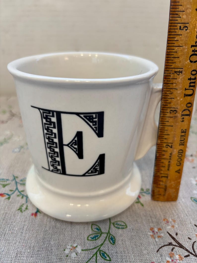 Puede incluir: Taza de cer&aacute;mica blanca con un dise&ntilde;o de la letra "E" en negro. La taza tiene un asa y una base ancha. Una regla de madera est&aacute; al lado de la taza, midiendo aproximadamente 10 cm de altura.