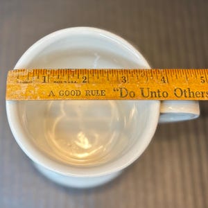 K&ouml;nnte beinhalten: Eine wei&szlig;e Keramiktasse mit Henkel, gemessen mit einem Holzlineal. Auf dem Lineal steht "A GOOD RULE 'Do Unto Others'". Der Durchmesser der Tasse betr&auml;gt ca. 7,6 cm.