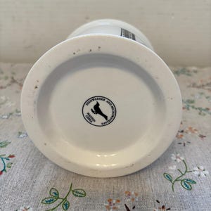 Puede incluir: Taza de cer&aacute;mica blanca con base ancha y parte superior ligeramente acampanada. La parte inferior de la taza presenta un sello en blanco y negro que dice "Apto para lavavajillas y microondas" y "Hecho en China".