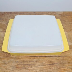 May include: A vintage yellow and white square container with a lid. The container has a bright yellow base and a white, translucent lid. The lid sits flush with the base, creating a clean, simple design. The container is likely made of plastic.
