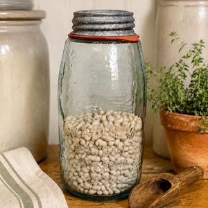 May include: A clear glass Mason jar filled with small white beans, featuring a textured surface and embossed lettering. The jar is topped with a metallic, ribbed screw-on lid and a thin red rubber gasket seal, creating a functional storage container.