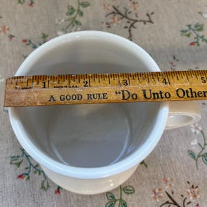 Puede incluir: Taza de cer&aacute;mica color crema con borde blanco, de aproximadamente 10 cm de di&aacute;metro, con asa. Una regla de madera est&aacute; colocada sobre la parte superior de la taza. La regla tiene el texto "A GOOD RULE 'Do Unto Others'" y "MADE IN U.S.A."