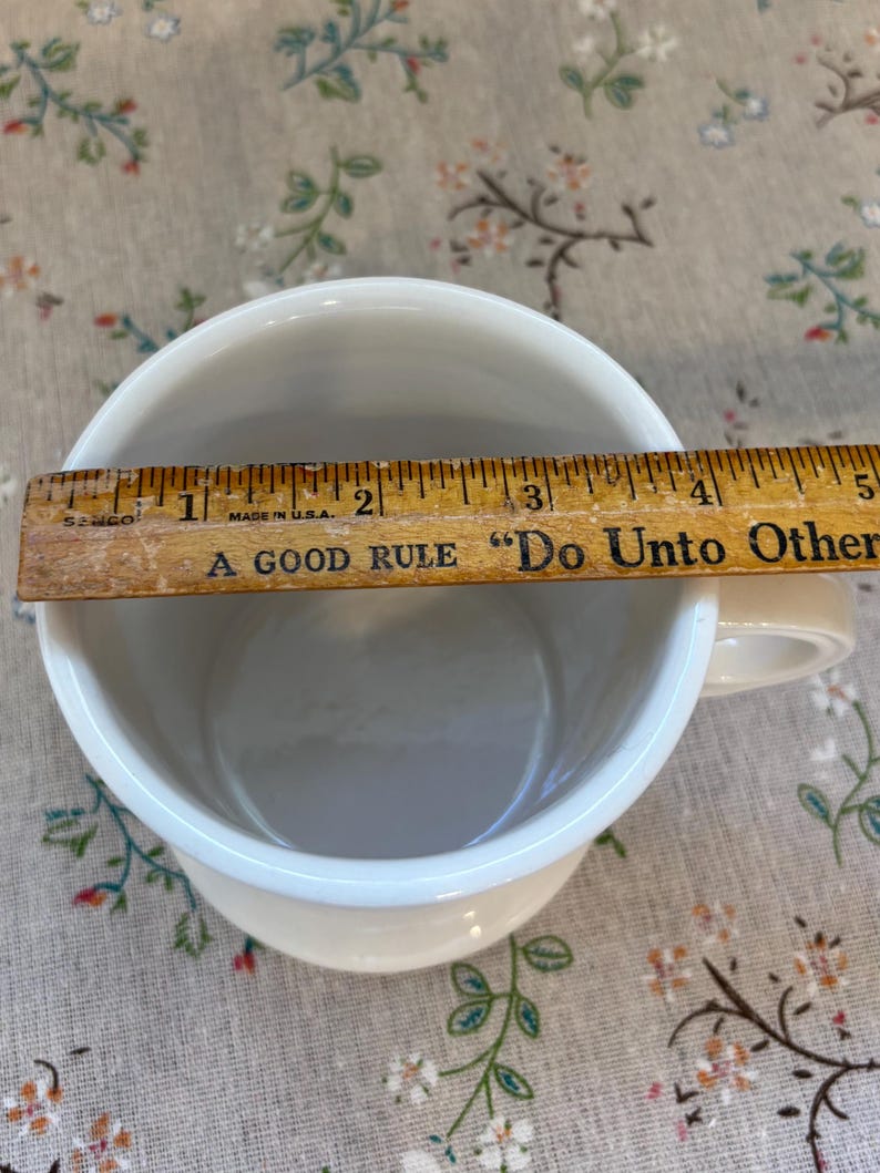 Puede incluir: Taza de cer&aacute;mica color crema con borde blanco, de aproximadamente 10 cm de di&aacute;metro, con asa. Una regla de madera est&aacute; colocada sobre la parte superior de la taza. La regla tiene el texto "A GOOD RULE 'Do Unto Others'" y "MADE IN U.S.A."