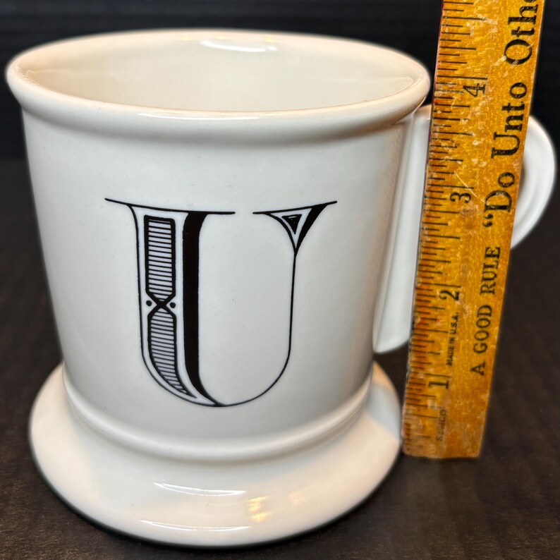 K&ouml;nnte beinhalten: Wei&szlig;e Keramiktasse mit einem schwarzen Buchstaben "U"-Design. Die Tasse hat einen Henkel und einen Sockel. Ein Holzlineal liegt neben der Tasse und misst etwa 10 cm.