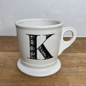 Pu&ograve; includere: Tazza in ceramica bianca con una lettera "K" nera. La tazza ha una base arrotondata e un manico. La lettera "K" &egrave; decorata con motivi decorativi. La tazza &egrave; su una superficie di legno.